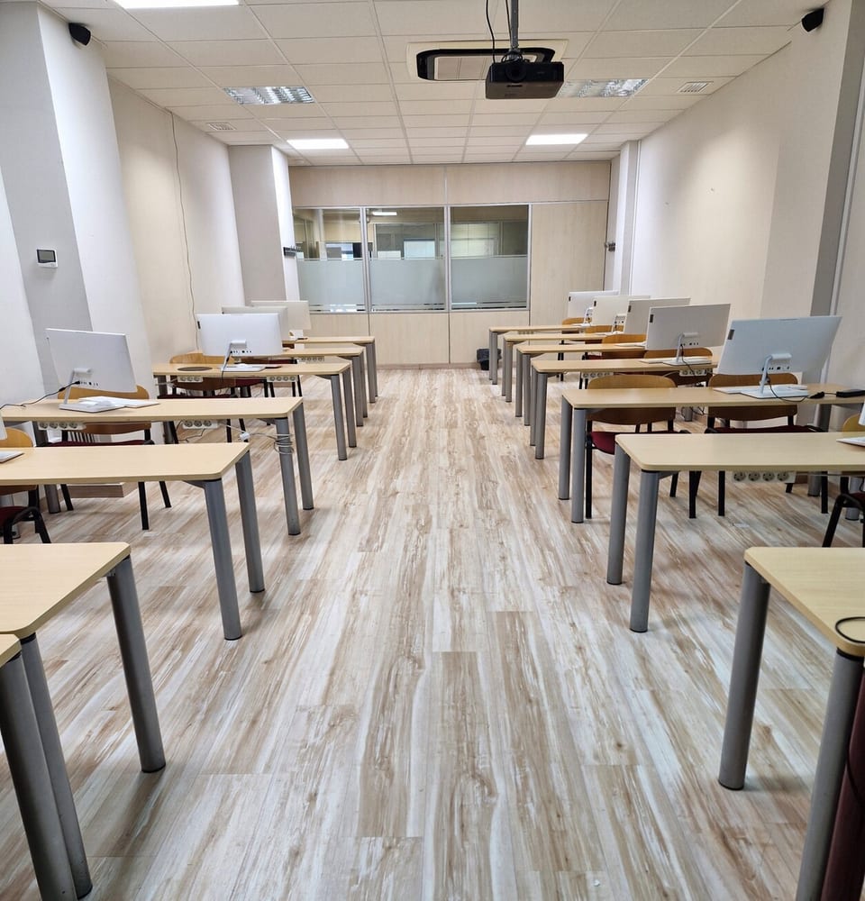 Aula de informática del centro Formación San Miguel en Zaragoza, equipada con puestos de ordenador all-in-one sobre mesas individuales de madera clara, proyector cenital, sala de cristal anexa al fondo y suelo de tarima vinílica
