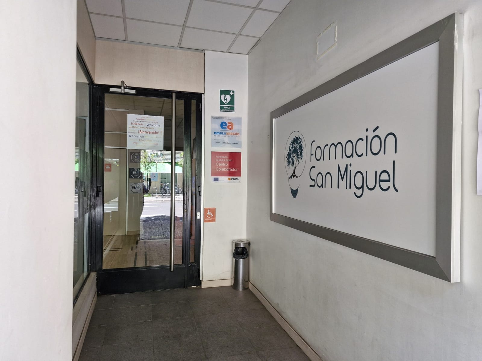 Entrada del centro Formación San Miguel en Zaragoza, con el logotipo corporativo (árbol del conocimiento dentro de una bombilla) rotulado en la pared, puerta acristalada de acceso con cartel de bienvenida en varios idiomas, y los distintivos de Centro Colaborador de Formación para el Empleo, agencia de colocación EmpleAragón, espacio cardioprotegido y accesibilidad