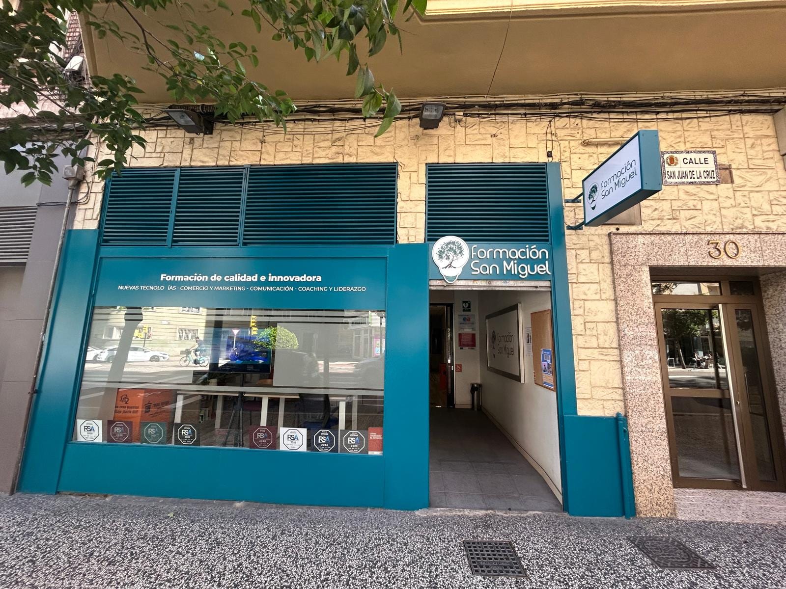 Fachada principal del centro Formación San Miguel en Calle San Juan de la Cruz 30 de Zaragoza, con rotulación corporativa "Formación San Miguel", isotipo grande de la bombilla con árbol del conocimiento en el cristal, y sello RSA Plus de Aragón Empresa pegado al escaparate