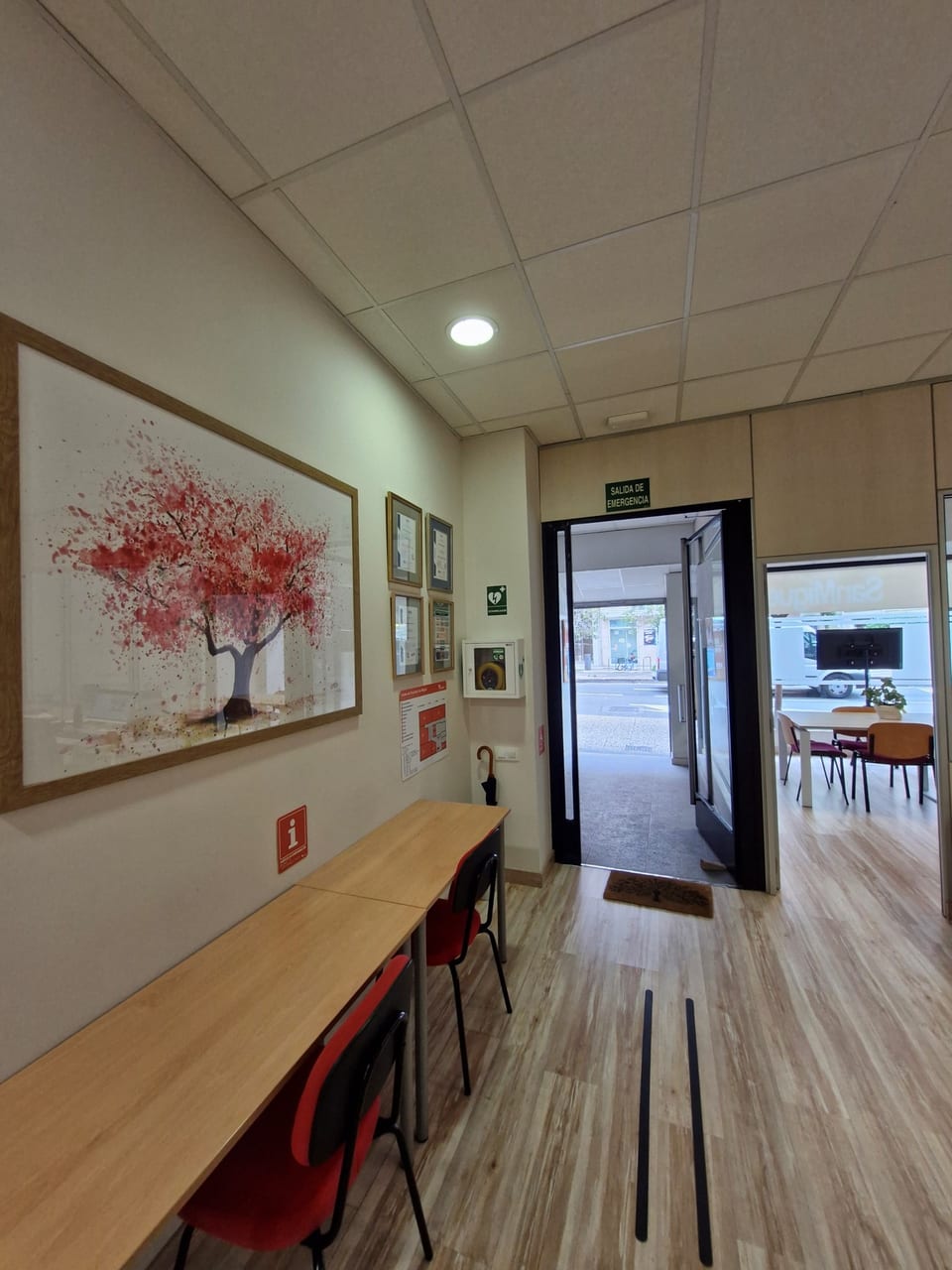 Hall de información del centro Formación San Miguel en Zaragoza, con desfibrilador AED, placas oficiales (Centro Colaborador INAEM, accesibilidad, salida de emergencia), zona de espera con mesa y sillas, cuadro decorativo y guías táctiles de accesibilidad en el suelo